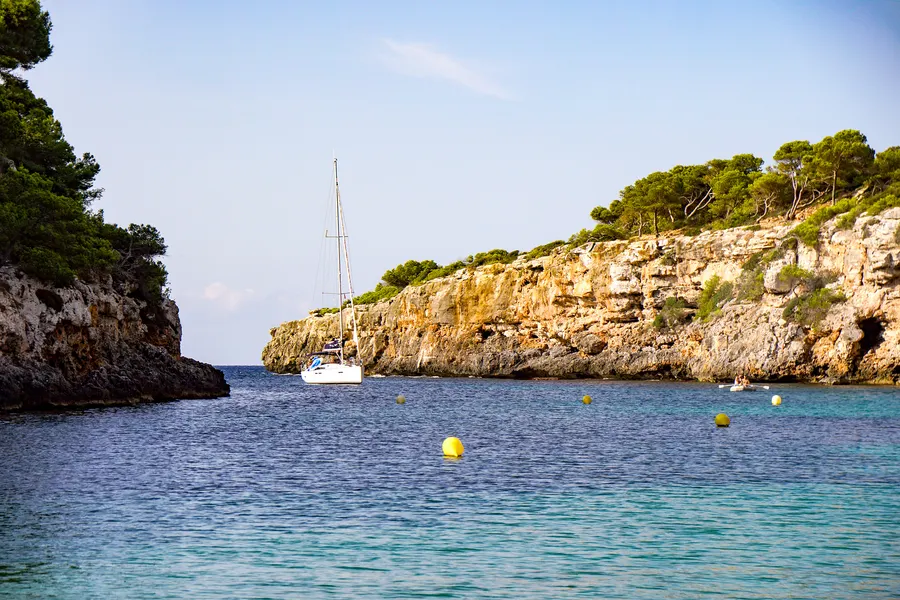 Ein weißes Segelboot ankert vor der Einfahrt von Cala Pi zwischen zwei Felswänden, im Vordergrund türkisfarbenes Wasser mit gelben Bojen.
