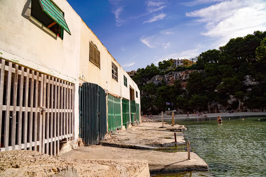 Die Reihe der Bootshäuser von Cala Pi mit grünen und hellen Lattentoren, davor die Slipbahn aus Beton, am Ende der Bucht der Strand mit Badegästen.
