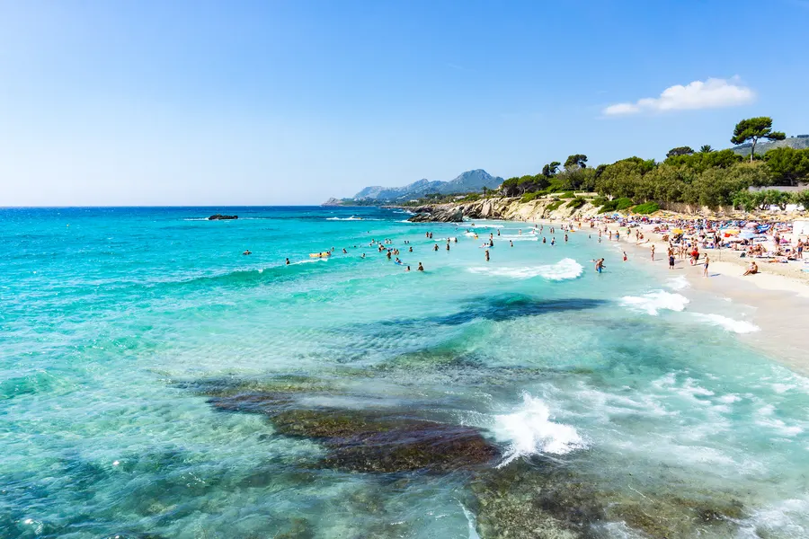 Der Ortsstrand Son Moll in Cala Ratjada: weite Sandbucht mit türkisfarbenem, sehr klarem Wasser und Badenden, im Vordergrund flache Felsen im Flachwasser.
