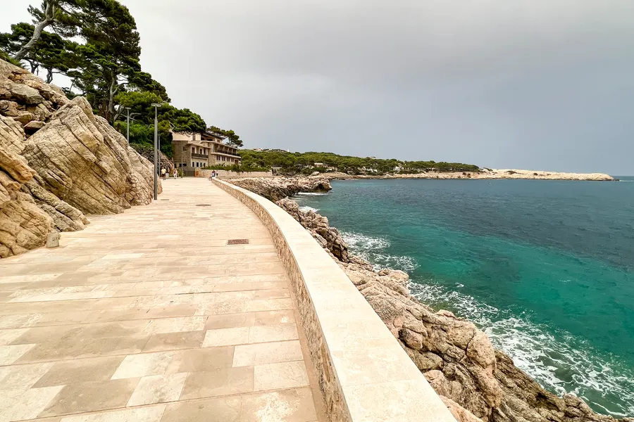 Gepflasterter Küstenweg, der über den Fels direkt am Wasser entlangführt, links Kiefern und Macchia, rechts das Meer.
