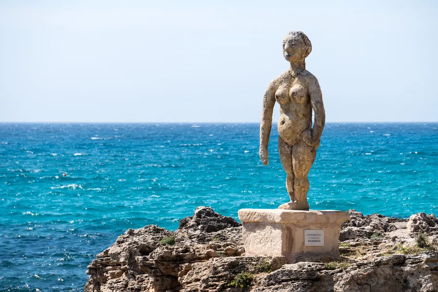 Bronzeskulptur einer stehenden Figur auf einem Steinsockel unmittelbar über der Felsküste, dahinter tiefblaues Meer.
