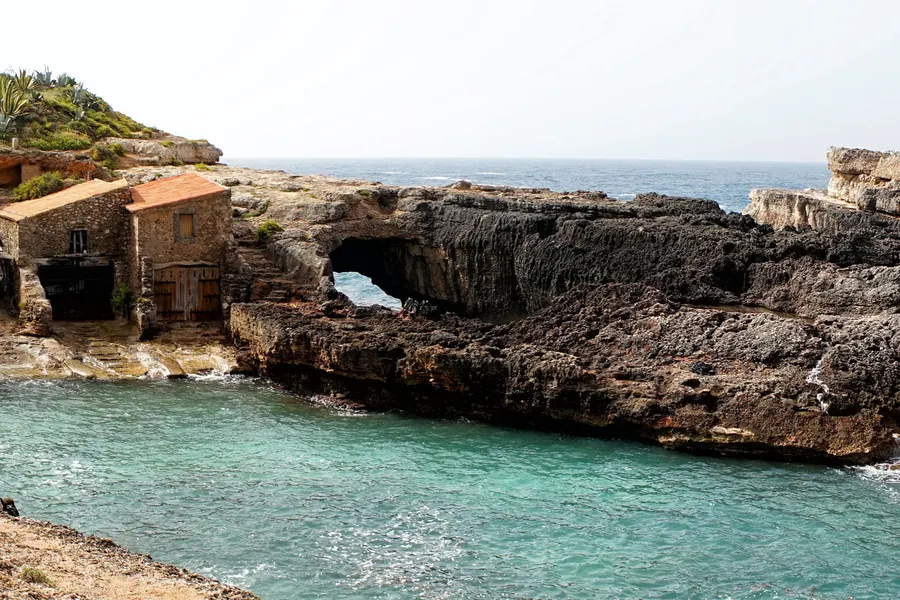 Blick über die felsige Cala s'Almunia mit sehr klarem türkisfarbenem Wasser, einzelnen Badenden auf dem Fels und niedrigem Bewuchs an den Rändern.
