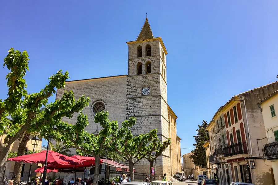 Blick vom Dorfplatz auf den Glockenturm der Kirche von Campanet, davor beschnittene Bäume und die Markise eines Lokals.