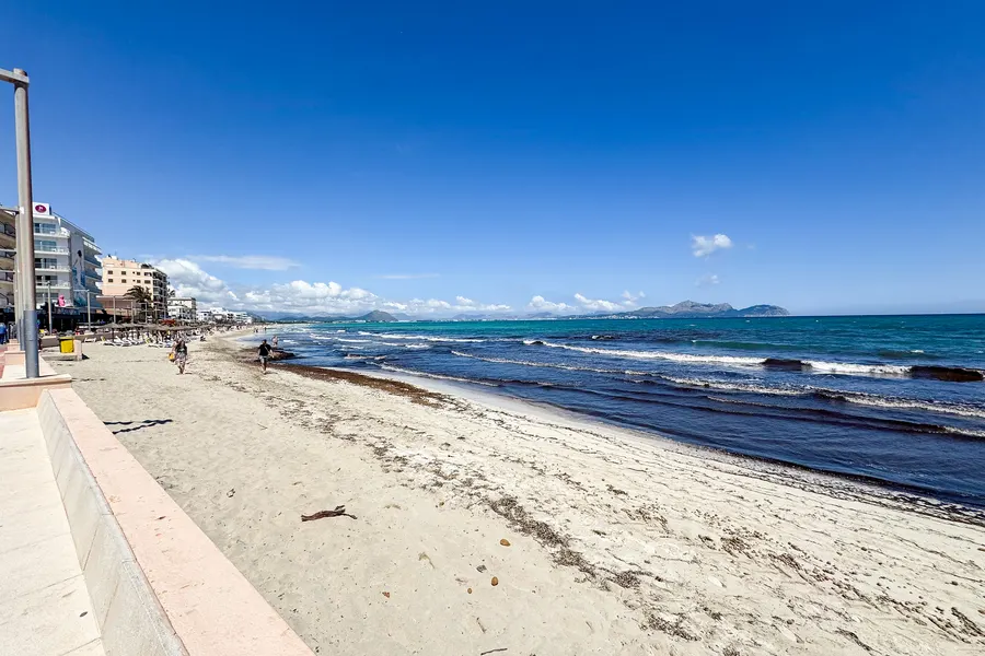 Derselbe Strandabschnitt im Frühjahr: breiter Sandstreifen mit ungeräumtem Seegrassaum am Wasser, vereinzelte Spaziergänger, links die Hotelzeile, am Horizont die Berge unter blauem Himmel.