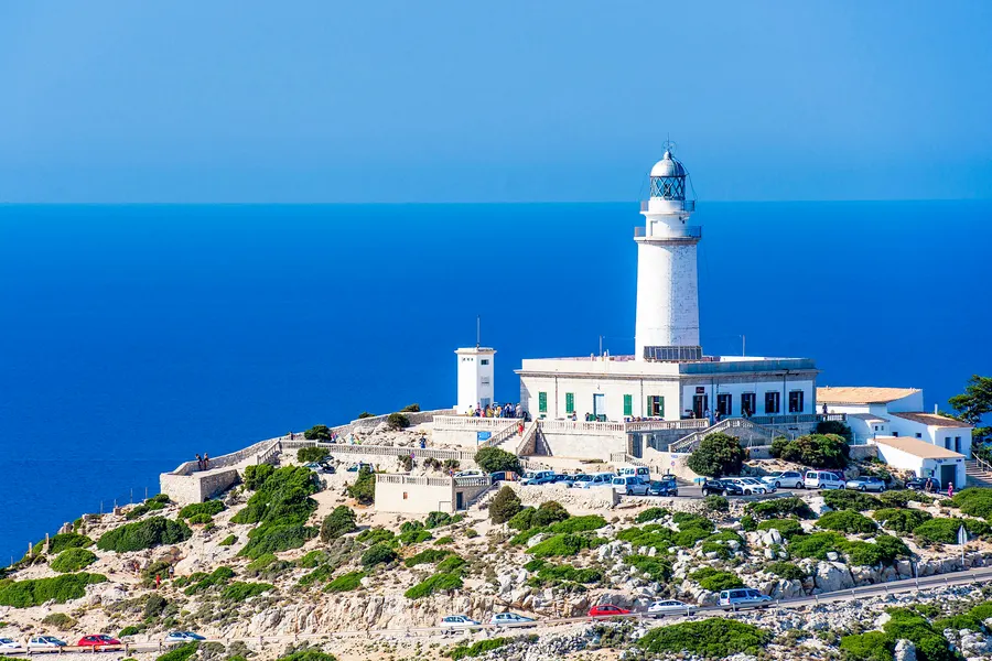 Der Leuchtturm von Formentor mit den umliegenden Gebäuden auf einer Felskuppe, davor halb gefüllte Parkflächen, ringsum tiefblaues Meer.