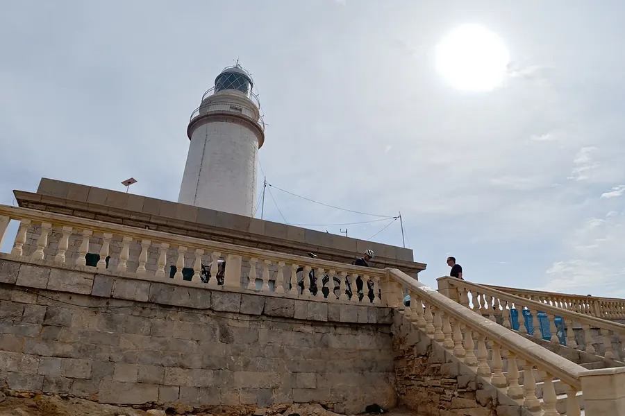 Steinerne Balustrade und Treppe hinauf zur Terrasse des Leuchtturms von Formentor, Menschen stehen am Geländer, dahinter der Turm im Gegenlicht.
