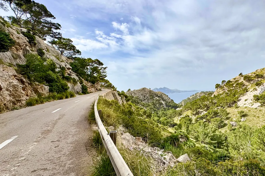 Schmale Bergstraße auf der Halbinsel Formentor, links eine Felswand mit Kiefern, rechts eine hölzerne Leitplanke und der Blick hinunter auf die Küste.
