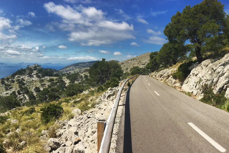 Die Straße zum Cap de Formentor führt am kahlen Hang entlang, links die Leitplanke über dem Abgrund, rechts Fels mit Kiefern.
