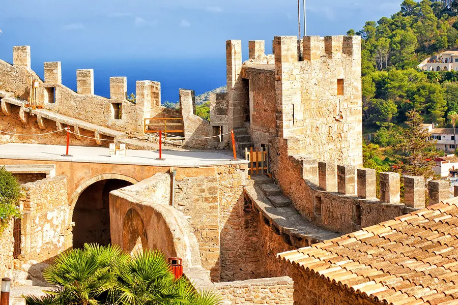 Begehbarer Wehrgang mit Zinnen im Castell de Capdepera, dahinter ein Turm und das flache Dach der Kapelle, im Hintergrund das Meer.