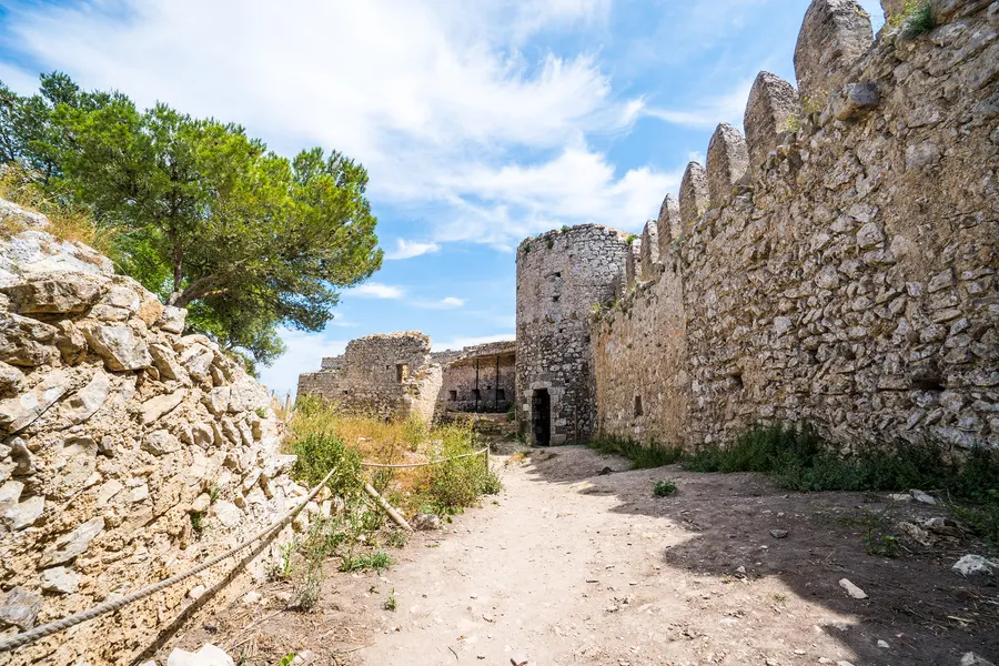 Unbefestigter Weg entlang der hohen Zinnenmauer des Castell de Santueri, links ein Kiefernbaum, rechts das Burgtor.