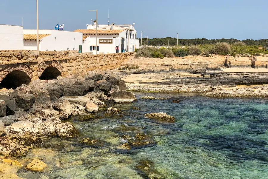 Flache zerklüftete Felsküste am Rand von Colònia de Sant Jordi mit klarem grünem Wasser und niedrigen weißen Häusern dahinter.
