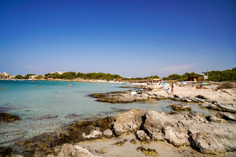 Flache, zerklüftete Felsplatten am Rand einer Badebucht bei Colònia de Sant Jordi, dahinter türkises flaches Wasser, Badende und ein Rettungsturm am Sandstrand.
