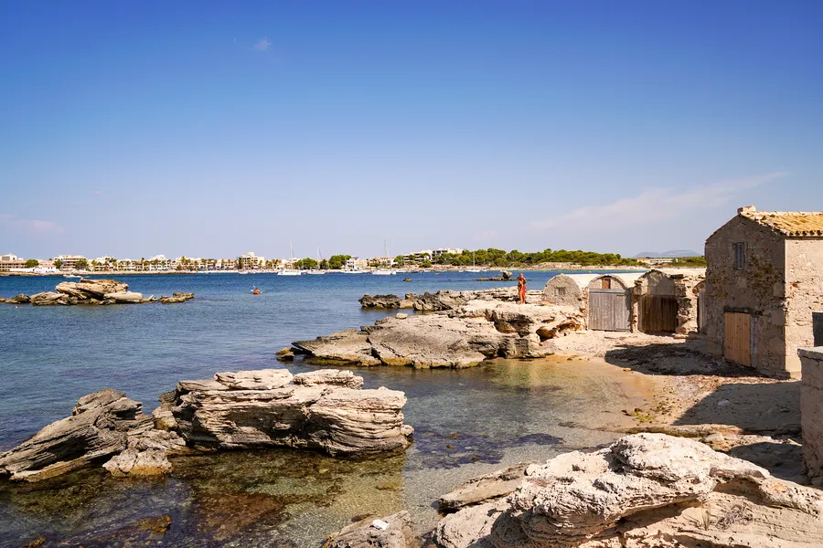 Zwei alte Häuser aus Naturstein mit Holztüren stehen unmittelbar an der flachen Felsküste bei Colònia de Sant Jordi, davor klares Wasser, am Horizont der Ort mit dem Hafen.
