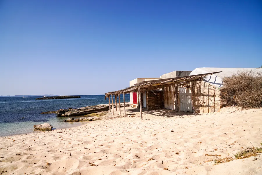 Ein niedriges Haus aus Naturstein mit vorgebautem Schilfdach steht direkt im Sand bei Colònia de Sant Jordi, links das flache Meer mit einem Felsriff.
