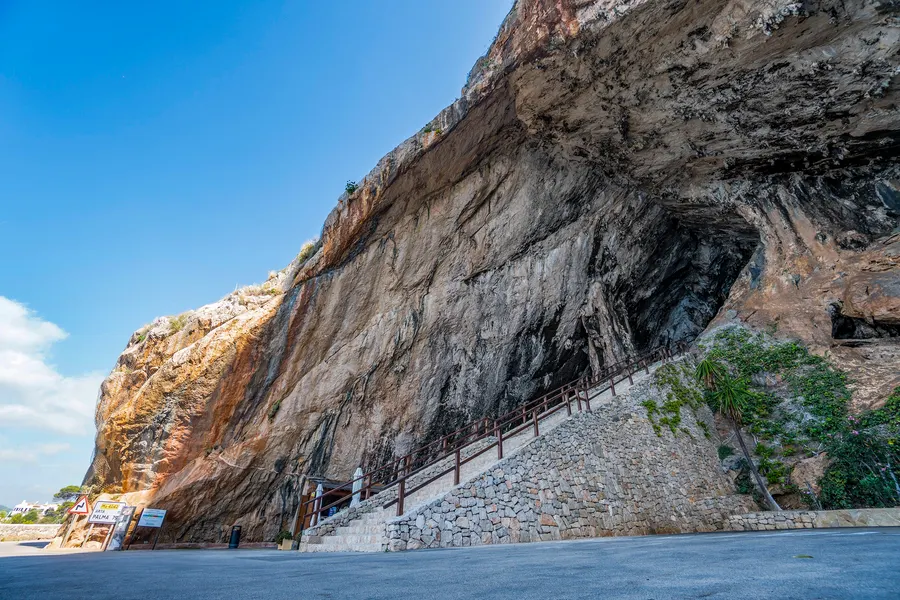 Hohe helle Felswand über dem Meer mit dem dunklen Höhlenportal und einer gemauerten Zugangstreppe, die an der Wand entlang hinaufführt.