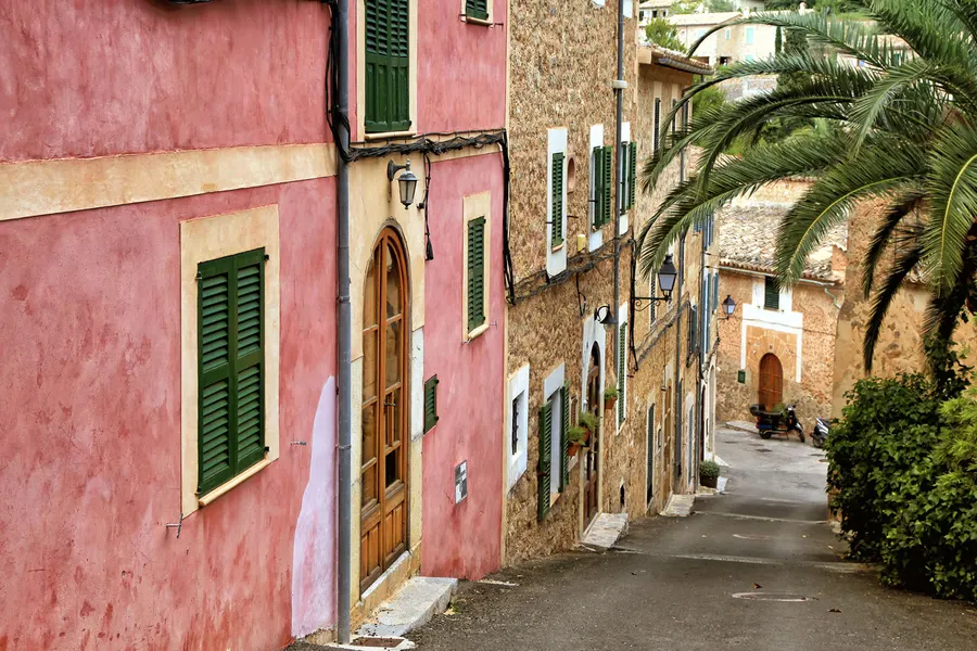 Gepflasterte Gasse in Deià mit einem rosa verputzten Haus, grünen Fensterläden und Natursteinmauern dahinter.
