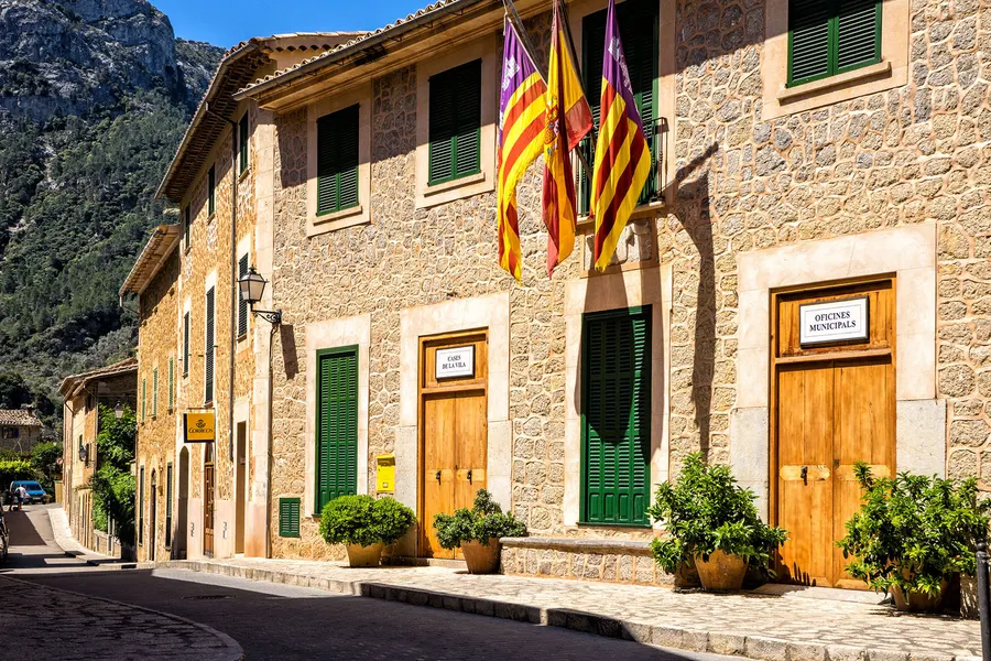 Das Rathaus von Deià aus hellem Naturstein mit grünen Fensterläden und den Fahnen Mallorcas und der Balearen über dem Eingang.
