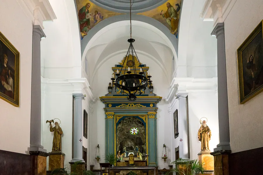 Innenraum der Kirche der Ermita de Betlem mit weißem Tonnengewölbe, Deckenfresken, einem blau-goldenen Altaraufsatz und zwei Heiligenfiguren auf dunklen Sockeln.
