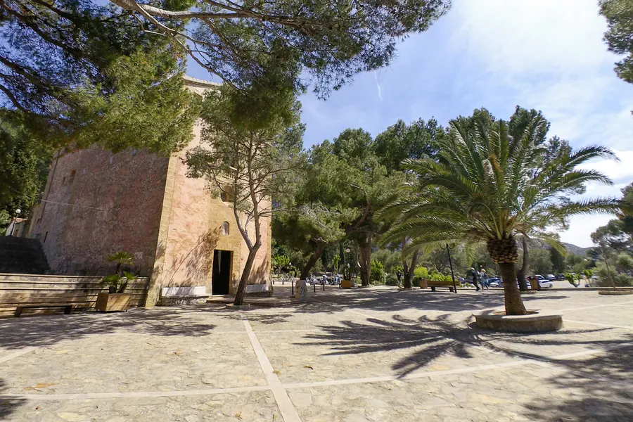 Der gepflasterte Vorplatz der Ermita de la Victòria unter hohen Kiefern und einer Palme, links die schlichte Steinfassade der Kirche, im Schatten stehen Bänke und geparkte Autos.
