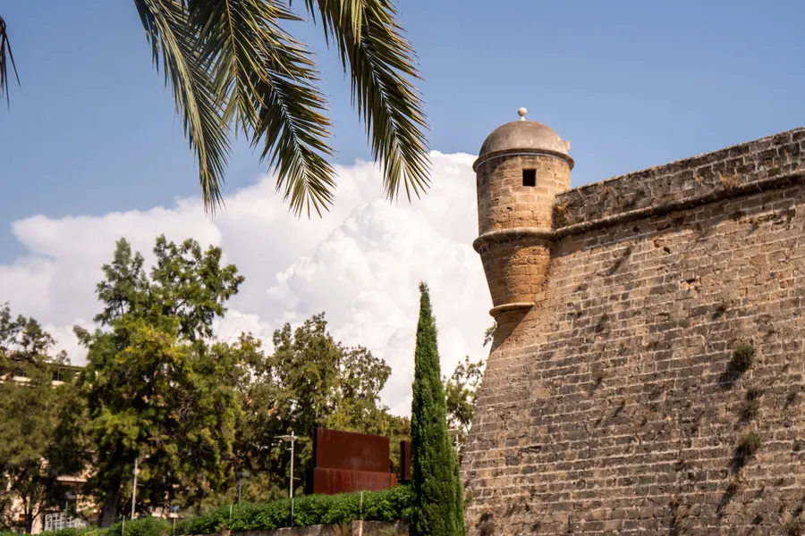 Runder steinerner Wachturm mit Kuppeldach auf der Ecke der Bastion Sant Pere in Palma, davor Zypressen und Palmen.

