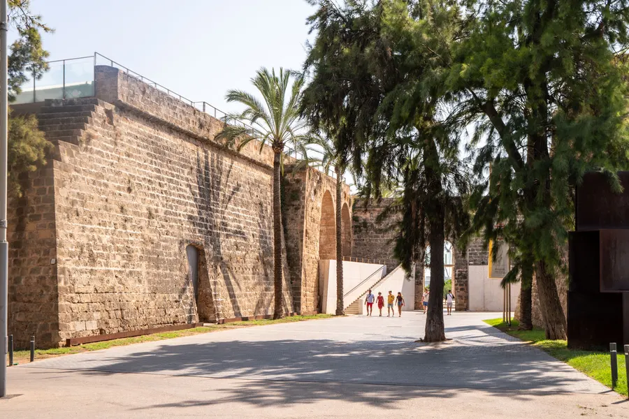 Breiter Weg entlang der hohen Bastionsmauer von Es Baluard in Palma, links Bäume, rechts eine Rasenfläche.
