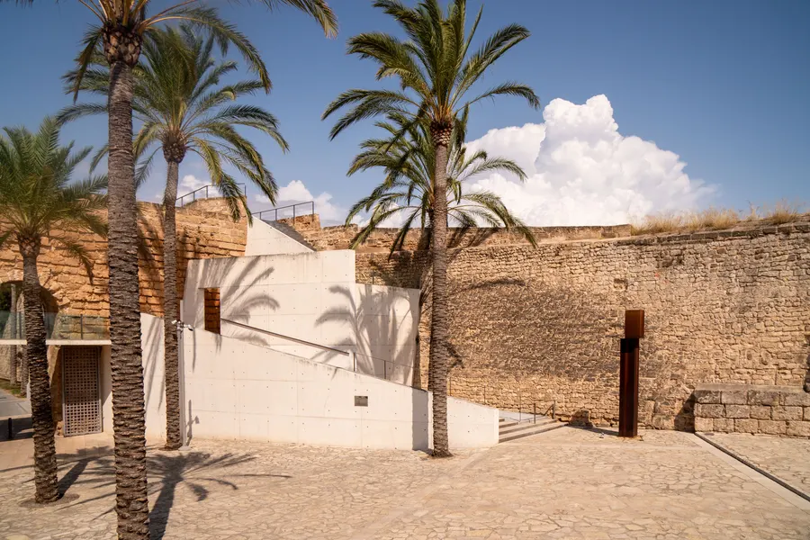 Offener Hof des Museums Es Baluard mit hohen Palmen, weißen Betonwänden und der alten Bastionsmauer im Hintergrund.
