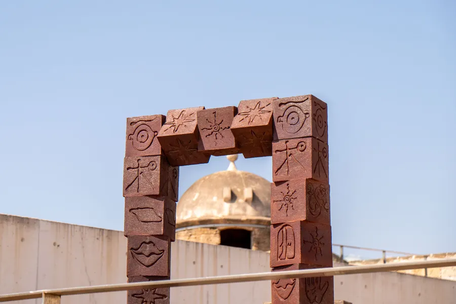 Rötliche Torskulptur mit eingeritzten Zeichen auf der Terrasse von Es Baluard, dahinter die Kuppel des alten Wachturms.