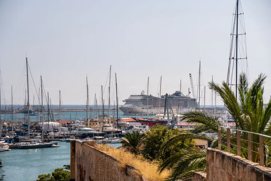 Blick von den Festungsmauern auf den Yachthafen von Palma mit Segelbooten und einem Kreuzfahrtschiff am Kai.
