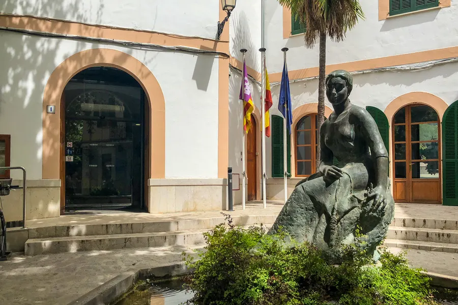 Rathaus von Esporles mit Bogenfenstern und Fahnen, davor ein Wasserbecken mit einer steinernen Figur und Grün.
