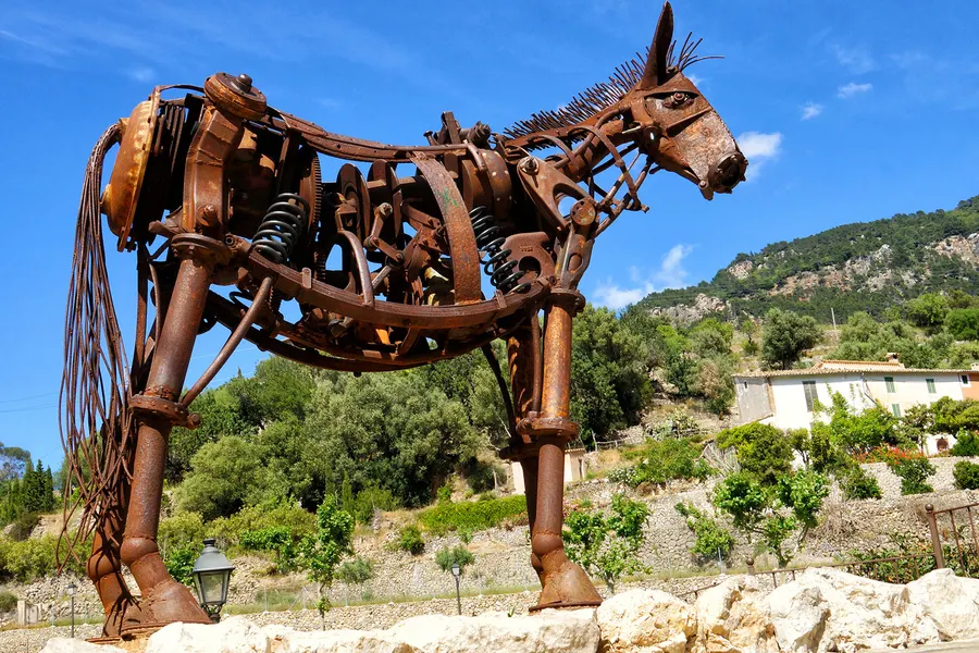 Lebensgroße Skulptur eines Maultiers aus verschweißten Alteisenteilen am Ortsrand von Estellencs, dahinter Terrassen.
