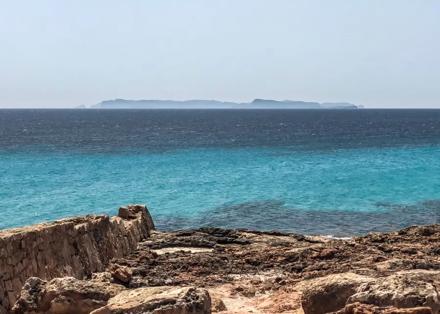 Blick über türkisfarbenes flaches Wasser und dunkelblaues Meer auf die lange flache Silhouette der Insel Cabrera am Horizont.