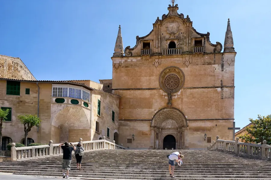 Die Kirche Sant Miquel in Felanitx mit barockem Giebel, Rundfenster und Portal, davor eine breite steinerne Freitreppe mit Balustrade und einzelnen Passanten.