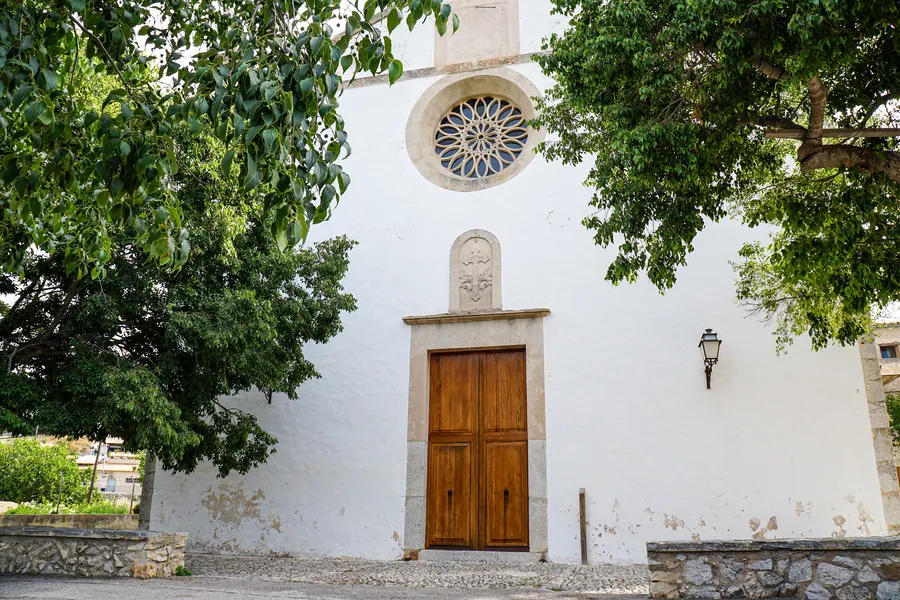 Die weiß getünchte Kirche von Galilea mit Rosettenfenster, hölzernem Portal und Wappenstein darüber, davor ein großer Baum.