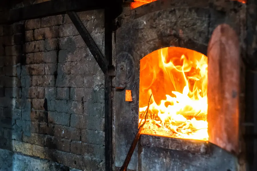 Offener Glasofen mit loderndem Feuer hinter einer Steineinfassung, davor eine Eisenstange im Glut.