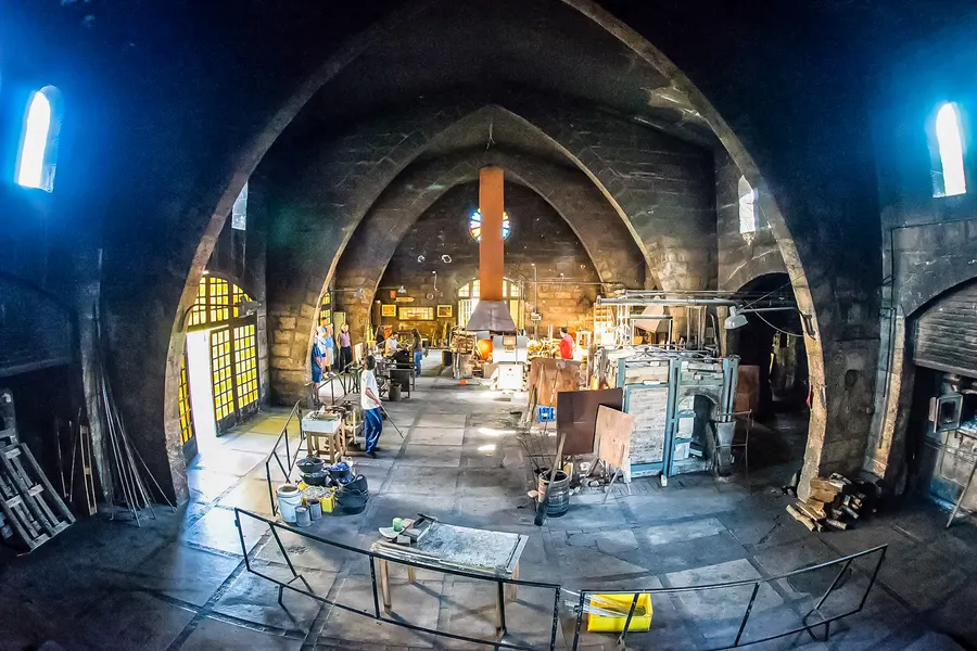 Große gewölbte Werkstatthalle der Glasbläserei mit Arbeitsplätzen, Werkbänken und Handwerkern im Hintergrund.