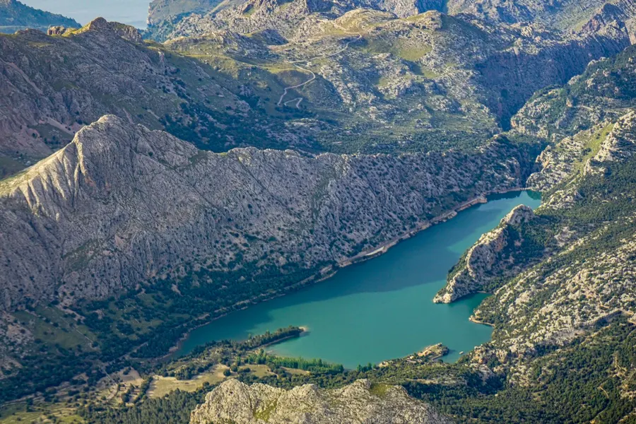 Luftaufnahme des Gorg Blau: der türkisgrüne Stausee liegt lang gestreckt in einem Tal der Serra de Tramuntana, umgeben von grauen Kalkgraten und bewaldeten Hängen, am oberen Bildrand die Serpentinen der MA-10.
