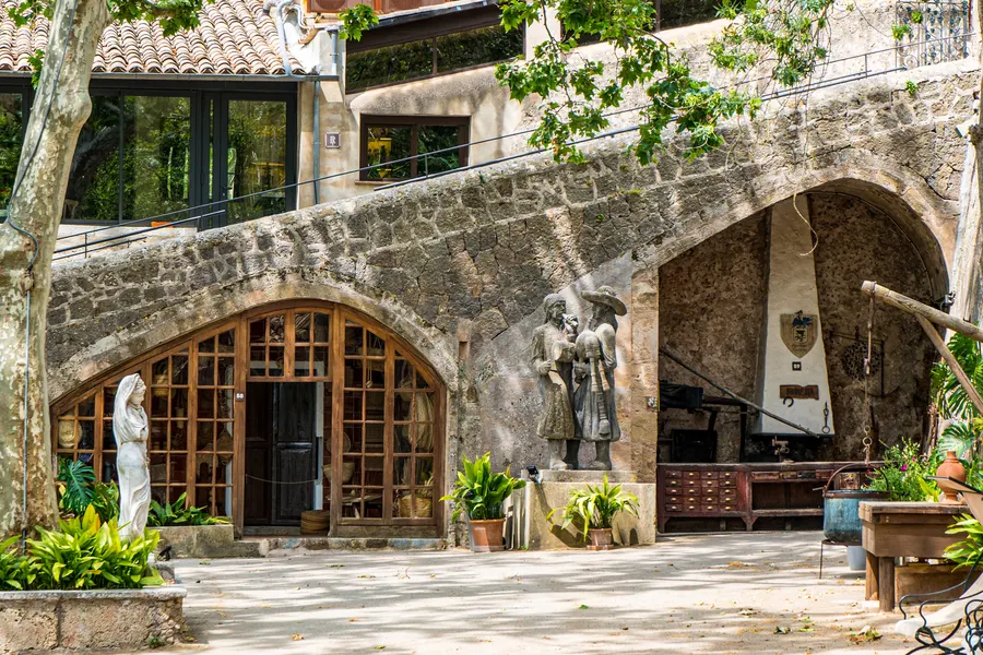 Gemauerter Bogengang im Hof von La Granja mit Werkstätten dahinter, Figuren, Werkzeug und Kübelpflanzen davor.