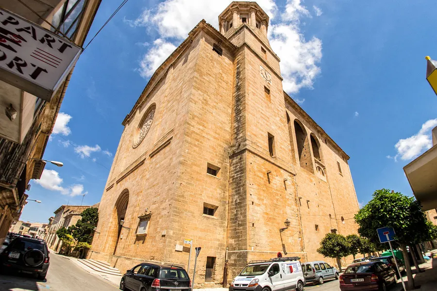 Die mächtige Pfarrkirche von Llucmajor aus hellem Sandstein mit Glockenturm, Uhr und Rundfenster, davor die Straße mit Autos.
