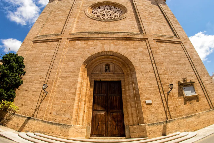 Die schlichte Sandsteinfassade der Kirche von Llucmajor mit Rundbogenportal und großem Rosettenfenster darüber.
