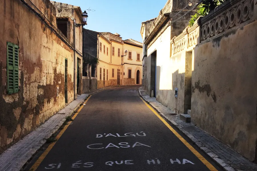 Abschüssige Gasse in Maria de la Salut zwischen einer hohen verputzten Mauer und Wohnhäusern, im Hintergrund die Ebene.
