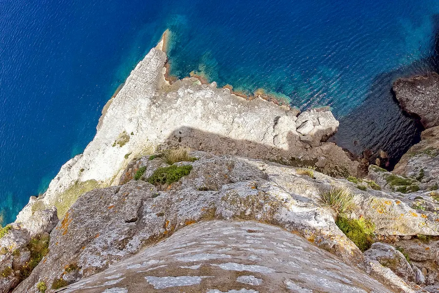 Senkrechter Tiefblick über die Felskante auf eine helle, spitz zulaufende Felsnadel im tiefblauen Wasser weit unten, mit türkis schimmerndem Flachwasser an ihrem Fuß.