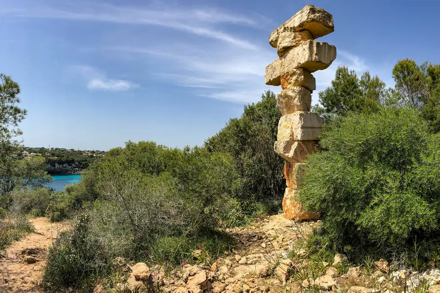 Aufgeschichtete Steinskulptur aus hellen Blöcken am Wegrand oberhalb der Küste, dahinter Macchia und das Meer.