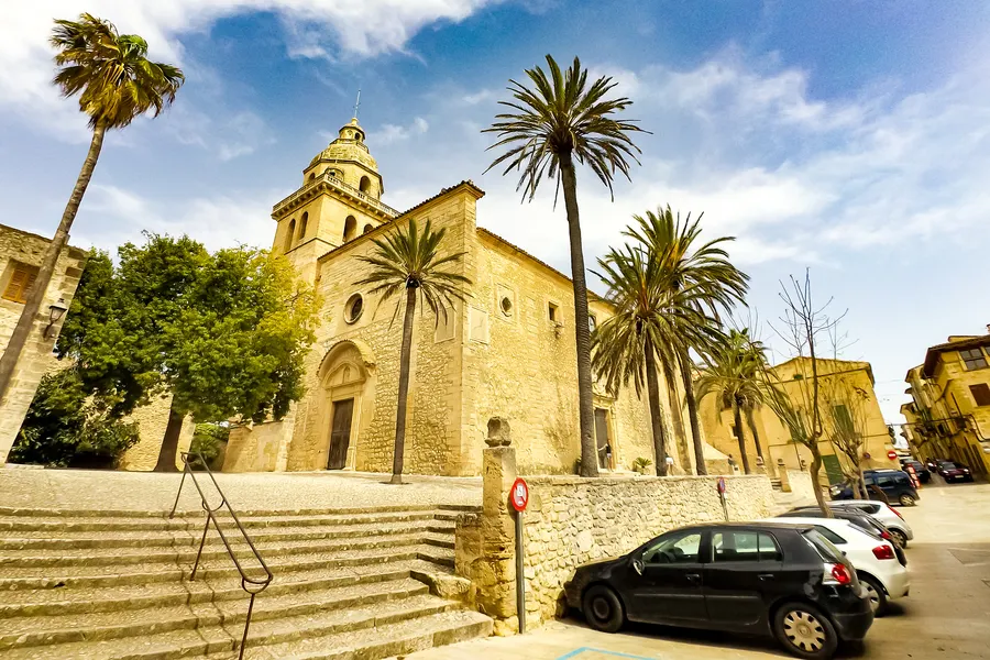 Die Pfarrkirche von Montuïri aus goldgelbem Sandstein mit Glockenturm und Freitreppe davor, umgeben von hohen Palmen unter bewölktem Himmel.