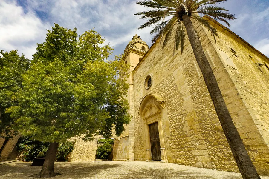 Die Fassade der Kirche von Montuïri aus goldgelbem Sandstein mit Spitzbogenportal, davor ein großer alter Baum und eine Palme.