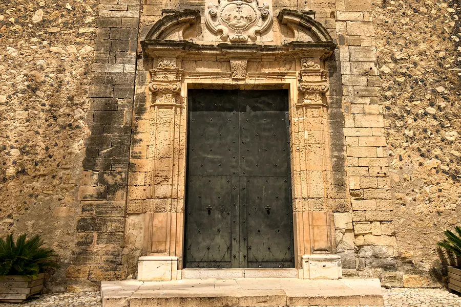 Reich verziertes Barockportal aus hellem Stein an einer groben Natursteinmauer, mit zweiflügeliger dunkler Holztür.
