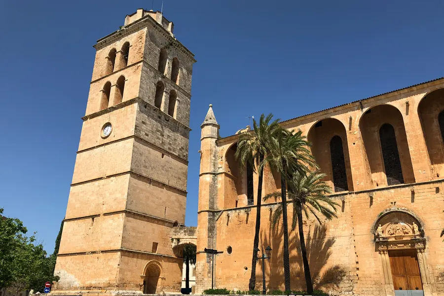 Die Kirche Sant Joan Baptista in Muro mit dem hohen freistehenden Glockenturm aus hellem Sandstein, daneben das Kirchenschiff mit Rundbogenarkaden und Palmen.