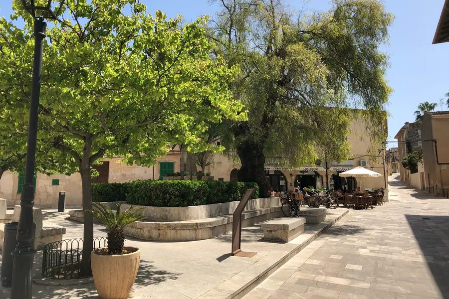 Schattiger Platz in Muro mit hohen Bäumen, Brunnen, Bänken und einer Bronzefigur, dahinter Häuser und Café-Terrassen.