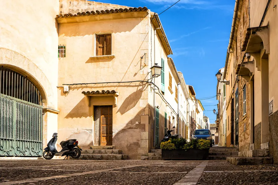 Gepflasterte Gasse in Muro mit Sandsteinhäusern, grünen Fensterläden, abgestellten Fahrrädern und Motorrollern.
