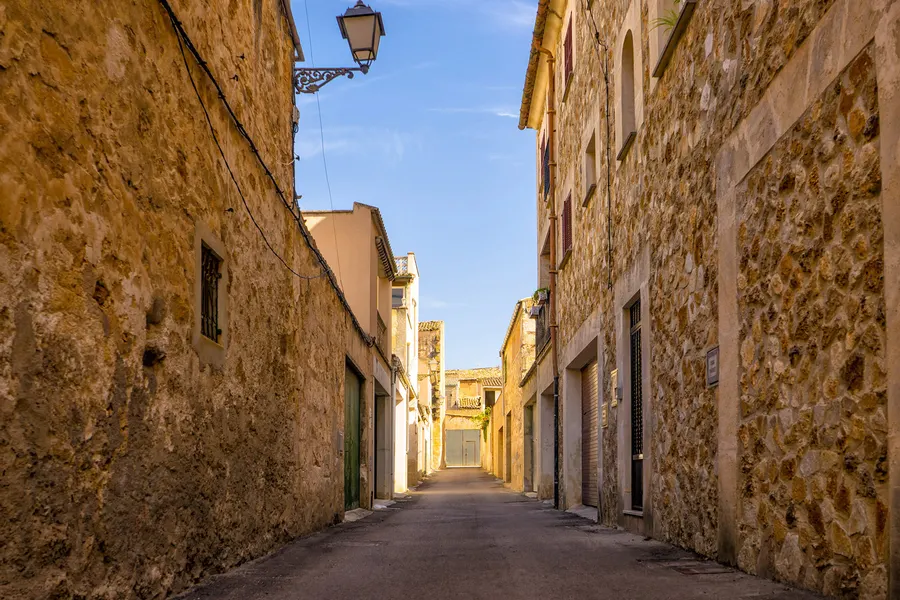 Enge Gasse zwischen hohen Sandsteinhäusern in Muro, am Ende der Kirchturm im Gegenlicht.
