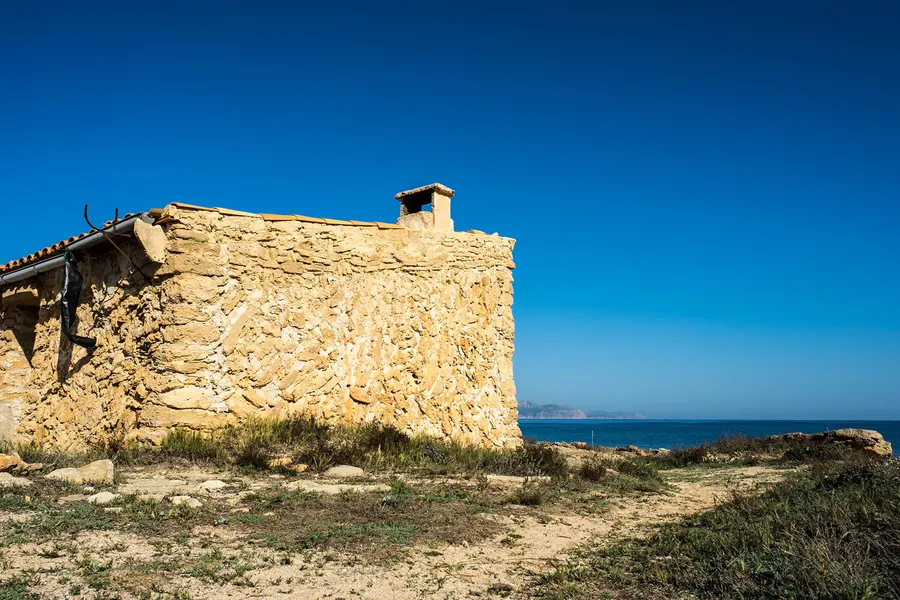 Altes Gebäude aus hellem Naturstein am Küstenweg bei Son Real, dahinter das Meer und die Bucht von Alcúdia.
